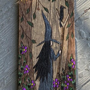 K&ouml;nnte beinhalten: Eine Holzschnitzerei einer Hexe, die in einem Wald mit einem Raben auf einem Ast steht. Die Hexe tr&auml;gt einen schwarzen Hut und ein schwarzes Kleid, und der Wald ist voller gr&uuml;ner Bl&auml;tter und lila Blumen. Die Schnitzerei ist etwa 15 cm hoch.