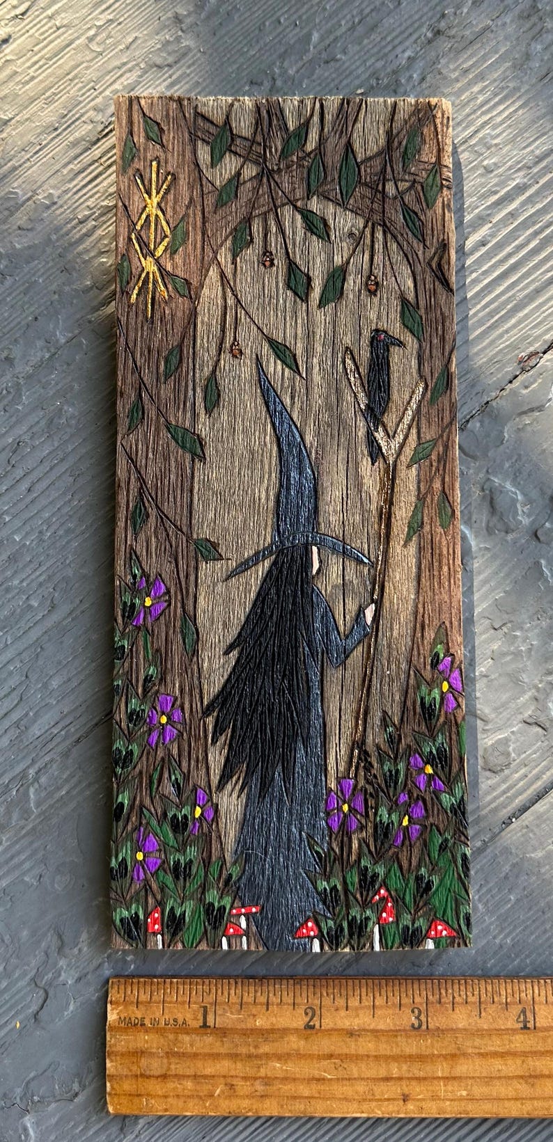 K&ouml;nnte beinhalten: Eine Holzschnitzerei einer Hexe, die in einem Wald mit einem Raben auf einem Ast steht. Die Hexe tr&auml;gt einen schwarzen Hut und ein schwarzes Kleid, und der Wald ist voller gr&uuml;ner Bl&auml;tter und lila Blumen. Die Schnitzerei ist etwa 15 cm hoch.
