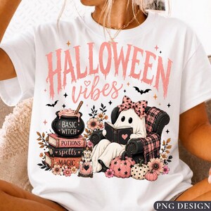 May include: A white t-shirt featuring a graphic with the text HALLOWEEN vibes in pink. The design includes a ghost wearing a pink bow reading a book in a chair, a cauldron labeled BASIC WITCH, stacked books labeled POTIONS, SPELLS, and MAGIC, and pumpkins.