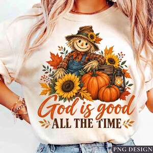 Peut inclure: Un t-shirt couleur crème avec une illustration d'un épouvantail portant un chapeau, assis à côté de deux citrouilles orange, d'une lanterne et de tournesols. Le texte "God is good ALL THE TIME" est imprimé en orange sous l'illustration.