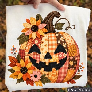 May include: A white t-shirt featuring a large, central pumpkin graphic composed of various orange, brown, and cream fabric patches with floral and gingham patterns. The pumpkin has a black felt jack-o'-lantern face, sunflowers, and autumn leaves.