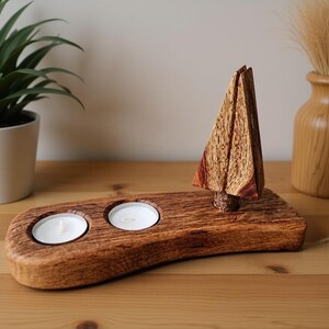 Könnte beinhalten: Ein hölzerner Kerzenhalter mit zwei brennenden Teelichtern und einem dekorativen Holzbaum. Der Kerzenhalter ist aus dunklem Holz gefertigt und hat ein natürliches, rustikales Aussehen. Im Hintergrund steht eine kleine Vase mit getrocknetem Gras.