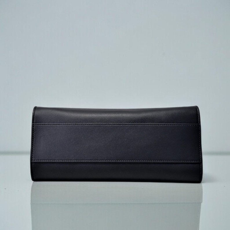 May include: A black leather clutch wallet with a rectangular shape. The wallet has a smooth texture with a horizontal seam detail across the center. The design is simple and elegant, suitable for various occasions.