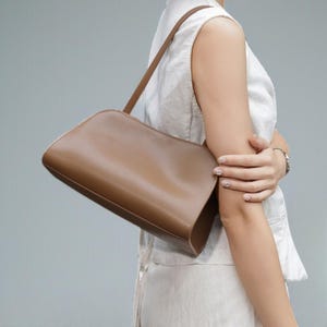 May include: A tan leather shoulder bag with a curved top and a long, thin strap. The bag is a solid color and appears to be made of smooth leather. The bag is being held by a person.