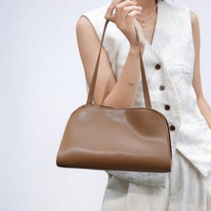 May include: A light brown, rectangular handbag with a curved top and long, thin straps. The bag is held by a person wearing a white vest. The bag's smooth surface and simple design suggest a minimalist aesthetic.