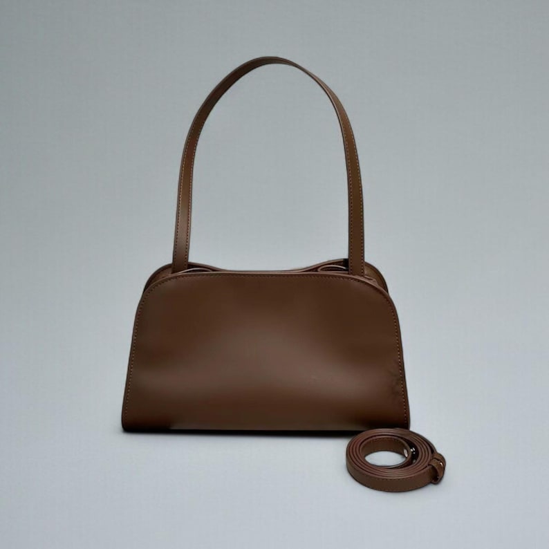 May include: A brown leather handbag with a curved top and two handles. A detachable shoulder strap is coiled beside the bag. The bag is a solid, medium brown color and appears to be made of smooth leather.