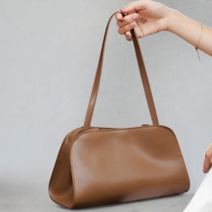 May include: A brown leather handbag with a rectangular shape and long, thin straps. The bag is held by a person's hand, showcasing its design and color against a neutral background. The bag's smooth texture and simple design suggest a classic style.
