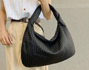 May include: A black woven shoulder bag with a large, curved shape. The bag has a thick, braided handle and a textured surface. The bag is held by a person wearing a striped shirt and tan pants.