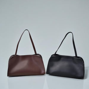 May include: Two shoulder bags are displayed. One is a deep brown color, and the other is black. Both bags have a rounded rectangular shape with a long, thin strap. The bags are made of a smooth material and are set against a light gray background.