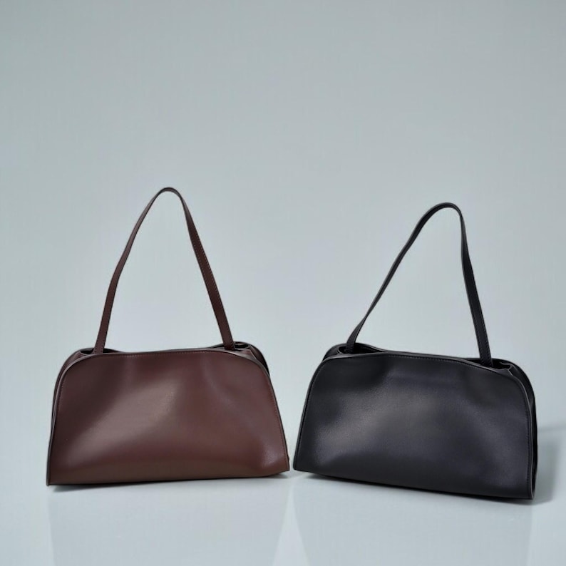 May include: Two shoulder bags are displayed. One is a deep brown color, and the other is black. Both bags have a rounded rectangular shape with a long, thin strap. The bags are made of a smooth material and are set against a light gray background.