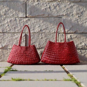 Può includere: Due borse tote rosse intrecciate con manici intrecciati. Le borse sono realizzate in un materiale intrecciato e hanno una forma trapezoidale. Le borse sono esposte su una superficie di pietra davanti a un muro di mattoni.