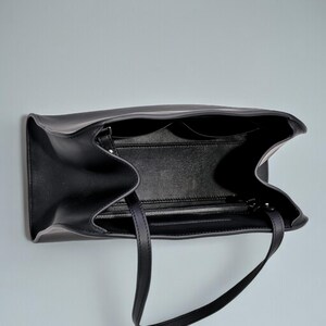 May include: Black leather tote bag with two handles, open to reveal interior pockets and a zippered compartment. The bag is shown against a light gray background, with a focus on its design and construction.