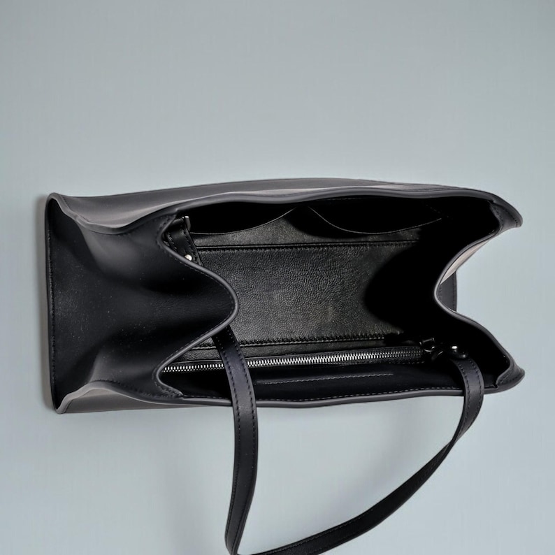 May include: Black leather tote bag with two handles, open to reveal interior pockets and a zippered compartment. The bag is shown against a light gray background, with a focus on its design and construction.