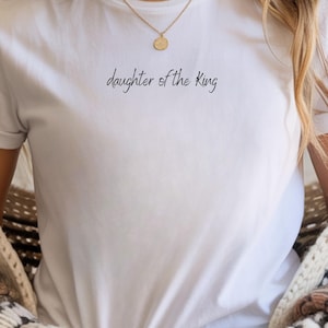 Könnte beinhalten: Weißes T-Shirt mit dem Schriftzug "daughter of the King" in schwarzer Schrift. Das Shirt hat einen Rundhalsausschnitt und kurze Ärmel. Eine goldene Halskette mit einem kleinen Anhänger ist zu sehen. Schlichtes Design.