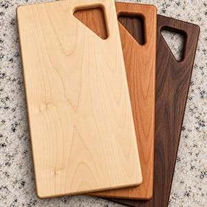 May include: Three rectangular wooden cutting boards stacked in a staggered arrangement. Each board features a smooth finish, rounded corners, and a triangular cutout handle at the top right corner. The boards are light, medium, and dark brown.