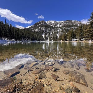 May include: A scenic landscape featuring a clear lake reflecting the surrounding trees and a snow-capped mountain under a bright blue sky. The foreground shows rocks and the lakebed. The scene evokes a sense of tranquility and natural beauty.