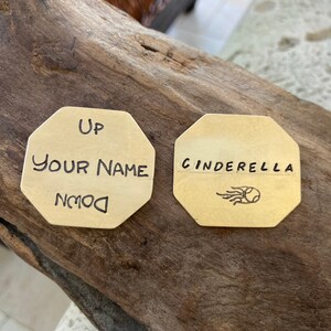 May include: Two octagonal brass tags with engraved text. The first tag says "Up Your Name Down" and the second tag says "Cinderella" with a small engraved image of a comet.