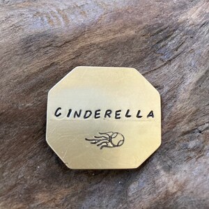 May include: A gold-colored metal tag with the word "CINDERELLA" engraved on it. A small image of a shooting star is below the word.