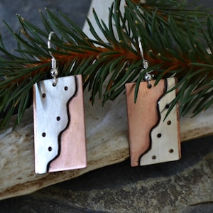 May include: A pair of silver and copper earrings with a wavy design and small silver dots. The earrings are rectangular in shape and have a silver hook.