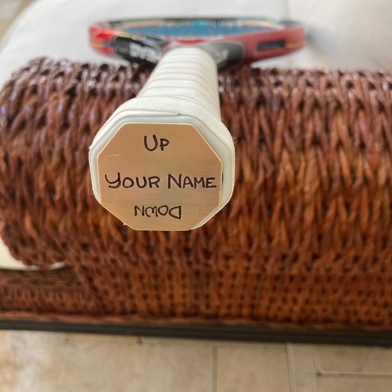 May include: A white tennis racket with a gold-colored octagonal plate on the handle. The plate has the words "Up", "Your Name", and "Down" engraved on it.