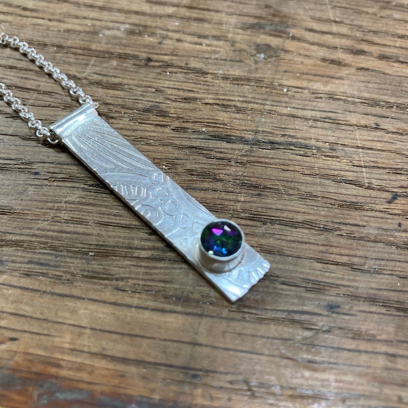 Pu&ograve; includere: Una collana con ciondolo in argento, una barra testurizzata e una pietra preziosa rotonda iridescente.