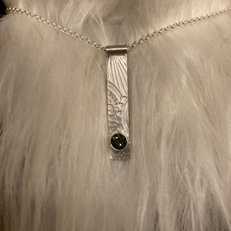 Pu&ograve; includere: Una collana d'argento con un ciondolo che presenta un design inciso dettagliato e una pietra preziosa verde.
