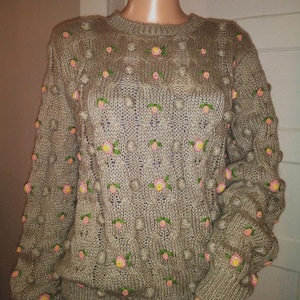 May include: A tan knit sweater with a crew neck and long sleeves. The sweater features a textured pattern with raised bobbles and is adorned with embroidered pink and yellow roses with green leaves. The sweater is a cozy and feminine garment.