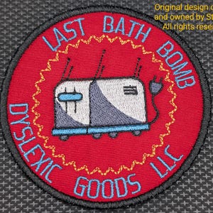 May include: Embroidered patch with a red background and a cartoon toaster design. The patch features the text "LAST BATH BOMB" and "DYSLEXIC GOODS LLC" in blue. The toaster is white and gray with blue accents. The design is surrounded by a yellow zig-zag border.