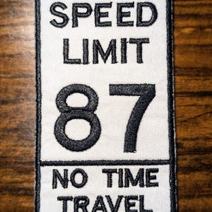 May include: A rectangular patch with a white background and black embroidered text. The text reads "SPEED LIMIT 87 NO TIME TRAVEL". The patch has a black border and is likely an iron-on or sew-on patch.