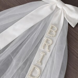K&ouml;nnte beinhalten: Ein wei&szlig;er Brautschleier mit einer gro&szlig;en Satinschleife oben. Ein wei&szlig;es Satinband mit dem Wort "BRIDE" aus perlenartigen Perlen ist am Schleier befestigt. Der Schleier besteht aus einem transparenten, leichten Stoff.