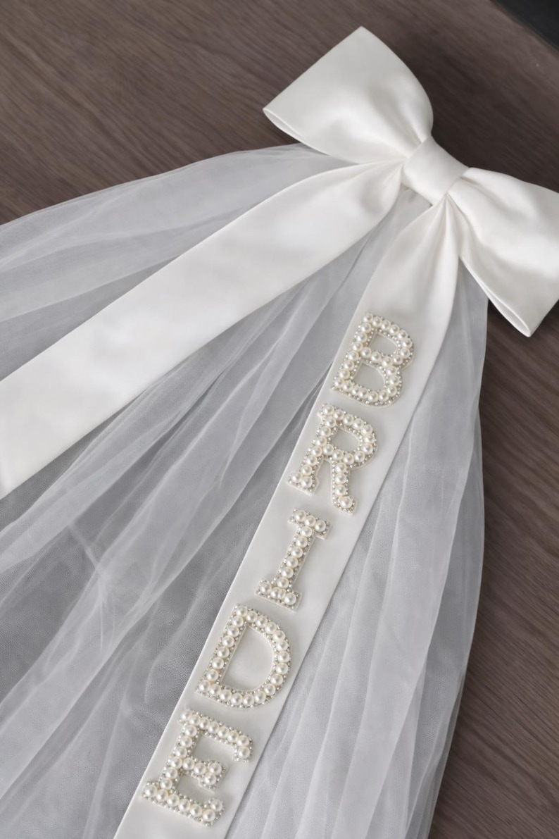 K&ouml;nnte beinhalten: Ein wei&szlig;er Brautschleier mit einer gro&szlig;en Satinschleife oben. Ein wei&szlig;es Satinband mit dem Wort "BRIDE" aus perlenartigen Perlen ist am Schleier befestigt. Der Schleier besteht aus einem transparenten, leichten Stoff.