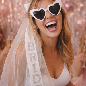 K&ouml;nnte beinhalten: Eine Braut tr&auml;gt eine wei&szlig;e Sonnenbrille in Herzform und einen Schleier mit dem Wort "BRIDE" in glitzernden Buchstaben. Sie l&auml;chelt breit, tr&auml;gt ein wei&szlig;es Kleid und eine wei&szlig;e Schleife im Haar. Rosa Ballons sind im Hintergrund zu sehen.