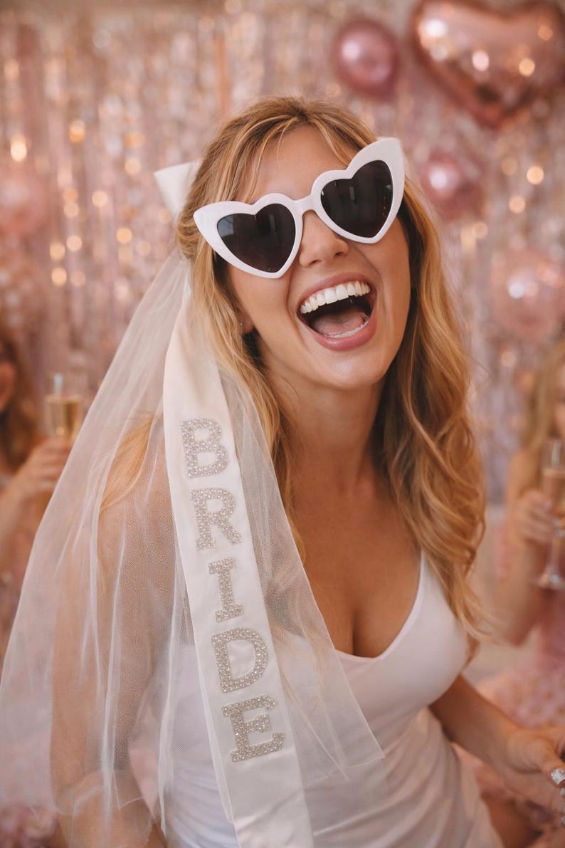 K&ouml;nnte beinhalten: Eine Braut tr&auml;gt eine wei&szlig;e Sonnenbrille in Herzform und einen Schleier mit dem Wort "BRIDE" in glitzernden Buchstaben. Sie l&auml;chelt breit, tr&auml;gt ein wei&szlig;es Kleid und eine wei&szlig;e Schleife im Haar. Rosa Ballons sind im Hintergrund zu sehen.