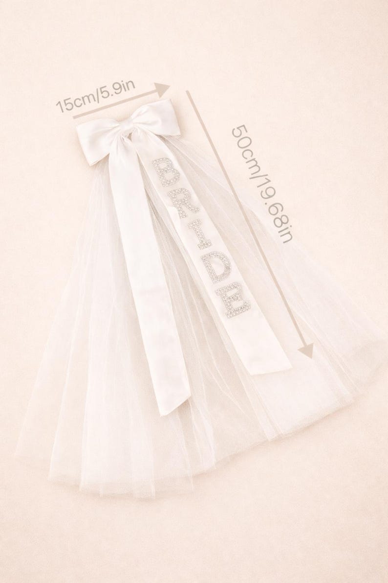 K&ouml;nnte beinhalten: Ein wei&szlig;er Brautschleier mit einer Satinschleife (15 cm) und einem Band mit der Aufschrift "BRIDE" in silbernen Strasssteinen. Der Schleier ist 50 cm lang. Der Schleier ist aus T&uuml;ll.