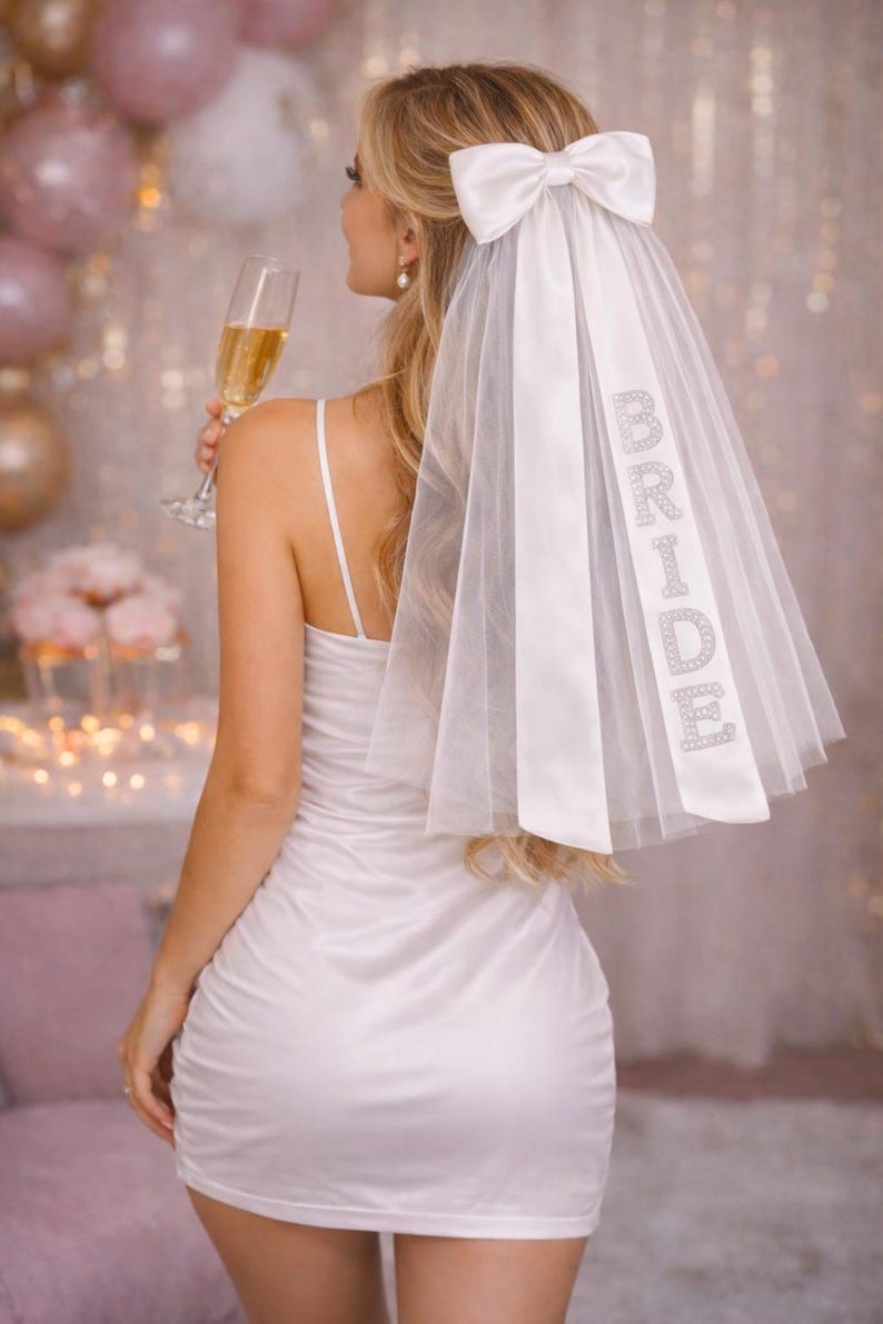 K&ouml;nnte beinhalten: Ein wei&szlig;er Brautschleier mit einer gro&szlig;en Schleife und dem Wort &bdquo;BRIDE&ldquo; in glitzernden Strasssteinen. Der Schleier ist an einem wei&szlig;en Satinband befestigt. Die Braut tr&auml;gt ein wei&szlig;es Kleid und h&auml;lt eine Champagnerfl&ouml;te.