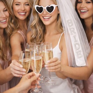 K&ouml;nnte beinhalten: Eine Gruppe von Frauen sto&szlig;t mit Champagner an. Eine Frau tr&auml;gt eine Sonnenbrille in Herzform und einen Schleier mit dem Text "Bride to Be". Die Frauen sind in Rosa- und Wei&szlig;t&ouml;nen gekleidet und feiern.