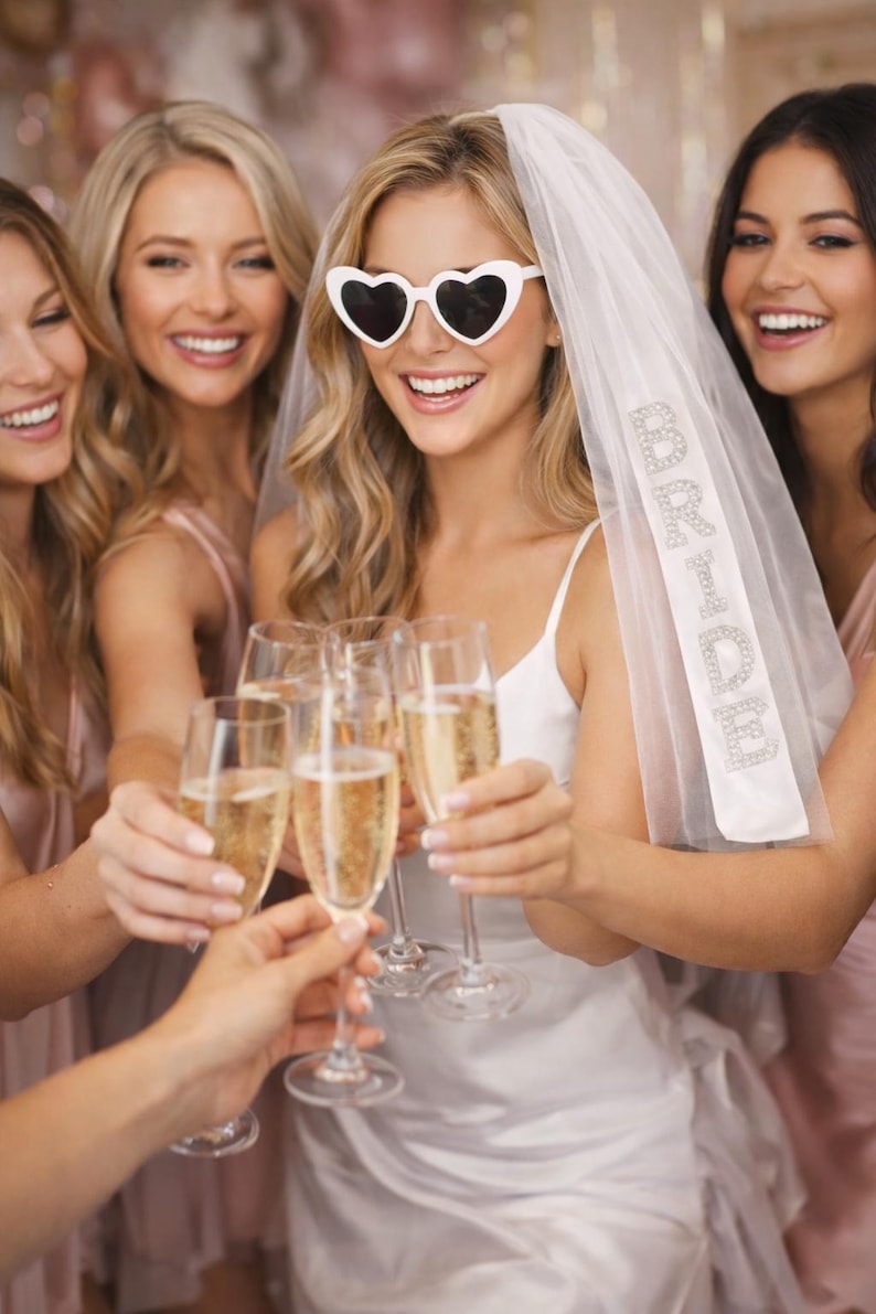 K&ouml;nnte beinhalten: Eine Gruppe von Frauen sto&szlig;t mit Champagner an. Eine Frau tr&auml;gt eine Sonnenbrille in Herzform und einen Schleier mit dem Text "Bride to Be". Die Frauen sind in Rosa- und Wei&szlig;t&ouml;nen gekleidet und feiern.