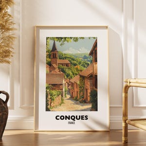 May include: A framed vertical art print featuring a colorful illustration of the village of Conques, France, with stone buildings, a church tower, and a cobblestone path. The text CONQUES FRANCE is printed in black at the bottom of the white border.