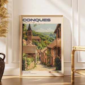 May include: A framed art print featuring a picturesque street scene in Conques, France. The artwork depicts stone buildings, a church steeple, and lush greenery. The print includes the text "CONQUES" and "FRANCE" with coordinates.