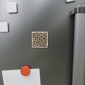 Puede incluir: Un imán cuadrado con un diseño de estampado de leopardo. El imán presenta un borde blanco y un patrón de manchas marrones sobre un fondo crema. El imán se muestra en un refrigerador gris.