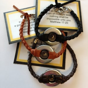 May include: Three leather bracelets with metal charms. Each bracelet has a different color of leather: black, brown, and tan. The charms are round and have a small seed inside. The bracelets have inspirational quotes on the cards they are attached to. The quotes are from the Bible, Matthew 17:20.