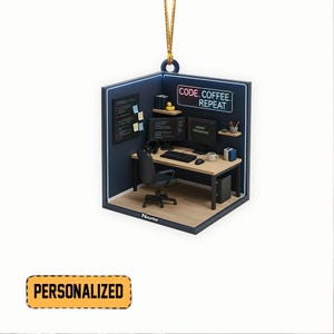 Pode incluir: Um enfeite personalizado que retrata um espa&ccedil;o de escrit&oacute;rio em miniatura com uma mesa, monitores de computador e uma cadeira de escrit&oacute;rio preta. O texto "CODE. COFFEE. REPEAT" &eacute; exibido acima dos monitores. Uma etiqueta amarela "PERSONALIZED" est&aacute; na parte inferior.