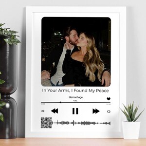 May include: A white framed print displaying a photograph of a couple kissing. The text "In Your Arms, I Found My Peace" is above a music player interface. The interface includes a waveform, song title, and artist.