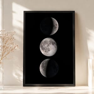 May include: A vertical rectangular black frame containing a print of three moon phases arranged in a column. The top moon is a waxing crescent, the middle is a full moon, and the bottom is a waning crescent, all rendered in shades of gray.