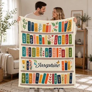 May include: A cream-colored throw blanket featuring a repeating pattern of horizontal shelves filled with colorful books and botanical illustrations. The name Josephine is printed in dark blue script in the center, surrounded by small butterflies.