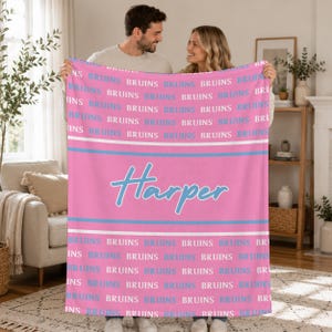 Könnte beinhalten: Eine rosafarbene Fleecedecke mit einem sich wiederholenden Muster des Wortes BRUINS in weißer und hellblauer Schrift, getrennt durch horizontale weiße und hellblaue Streifen. In der Mitte der Decke steht der Name Harper in hellblauer Schreibschrift.