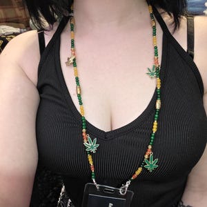 May include: A beaded necklace with green, yellow, and orange beads. The necklace features green marijuana leaf charms and gold-colored accents. A clear badge holder with the text "Mystic Alchemy" is also visible.