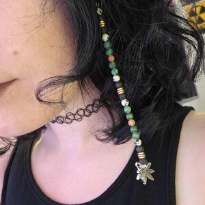 May include: A hair accessory featuring a strand of beads in green, white, orange, and black, accented with gold-colored spacers. The strand is adorned with a silver-colored marijuana leaf charm. The person is wearing a black tank top and a black choker.