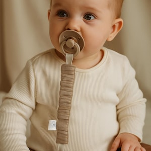 Può includere: Un bambino con un ciuccio beige e un fermaciuccio abbinato. Il fermaciuccio è realizzato in tessuto marrone chiaro strutturato e ha una perlina di legno all'estremità. Il bambino indossa una maglia a maniche lunghe a coste color crema e pantaloni marroni.