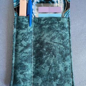 May include: A rectangular fabric pouch featuring a dark teal mottled texture on the front pocket and a blue and black agate-inspired print on the top flap. It contains a light pink highlighter, a blue ballpoint pen, and two adhesive note strips.